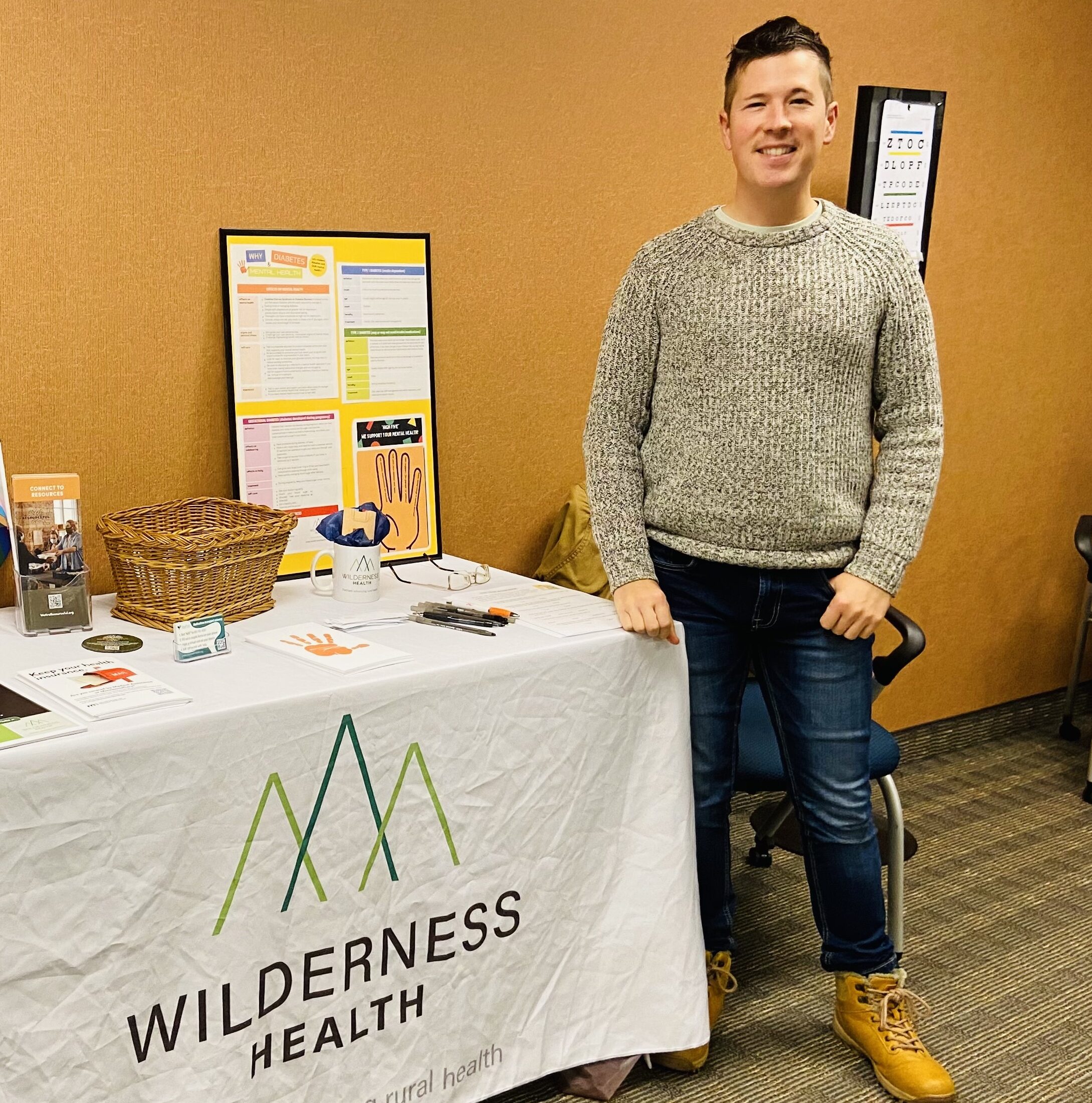 Smiling person stands next to a table with a tablecloth reading Wilderness Health.