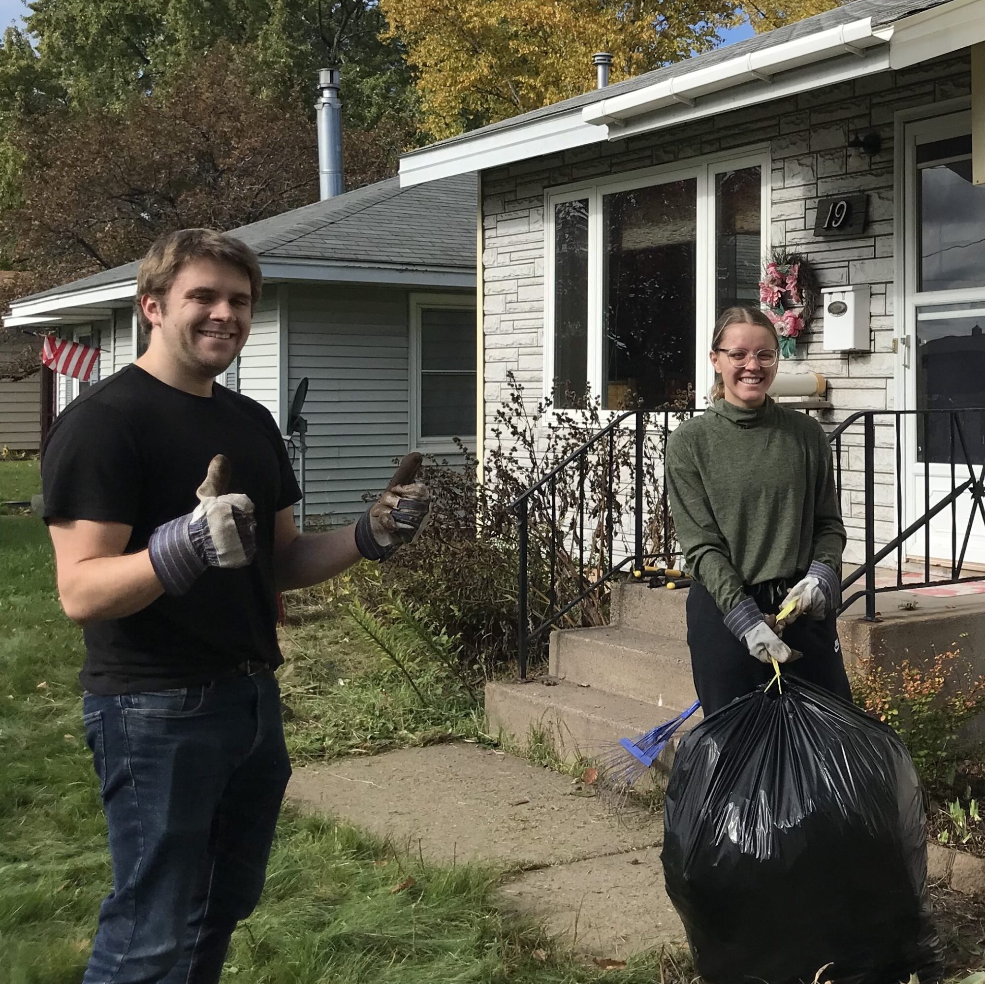 Two people wearing gardening gloves smile outside a home, one person is holding a full trash bag