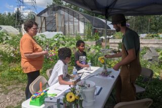 Person standing behind a table talks to a family standing across the table. They are outside and the table has flowers on it