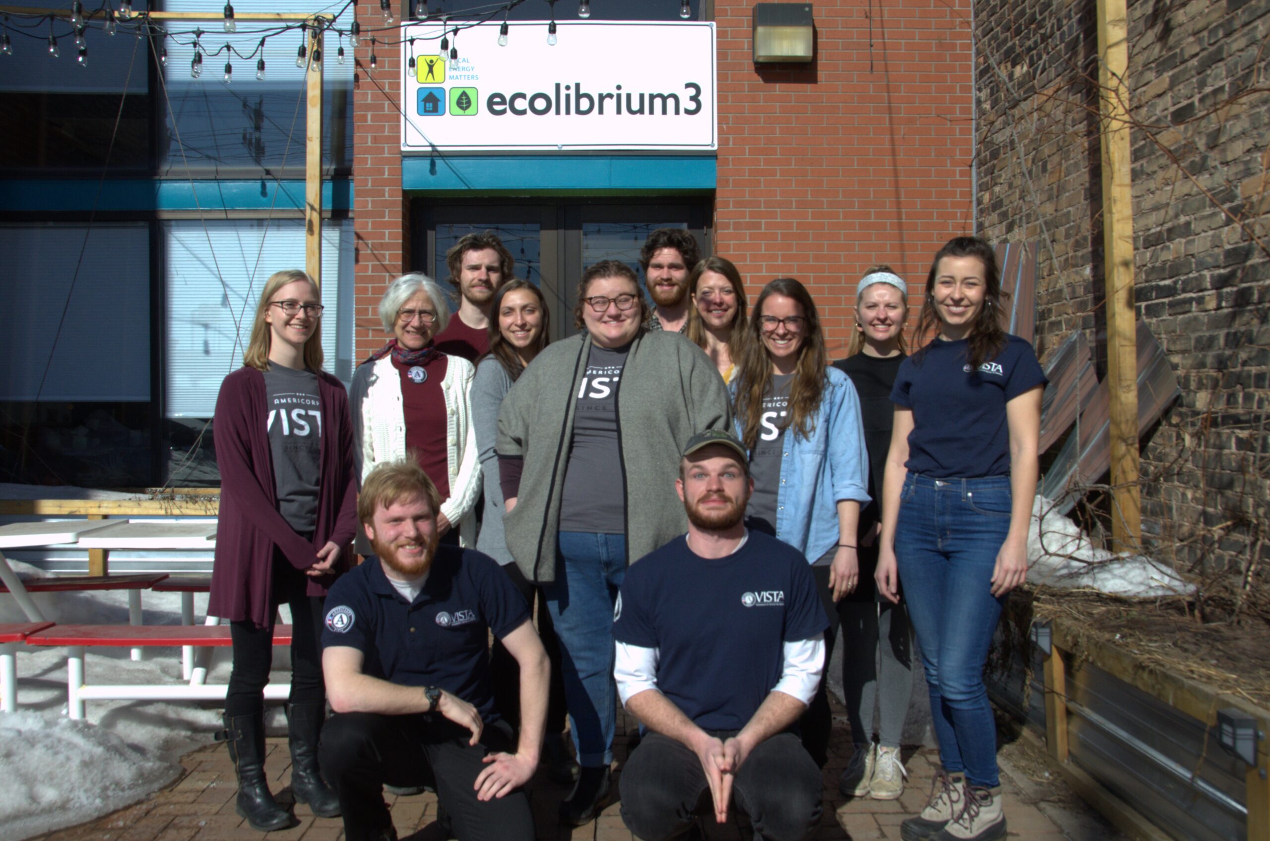 Twelve people pose for a group photo in front of a brick building with an Ecolibrium3 sign on it.