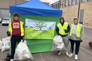 Three people wearing high-visibility vests and holding garbage bags smile and pose by an Earth Week banner.