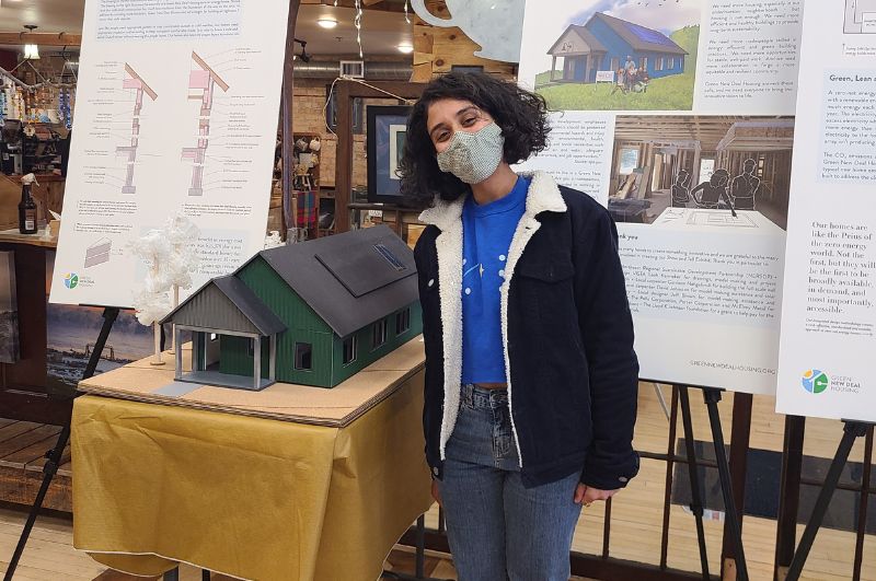 Person wearing a face mask smiles, standing in front of several display boards and a model of a house.