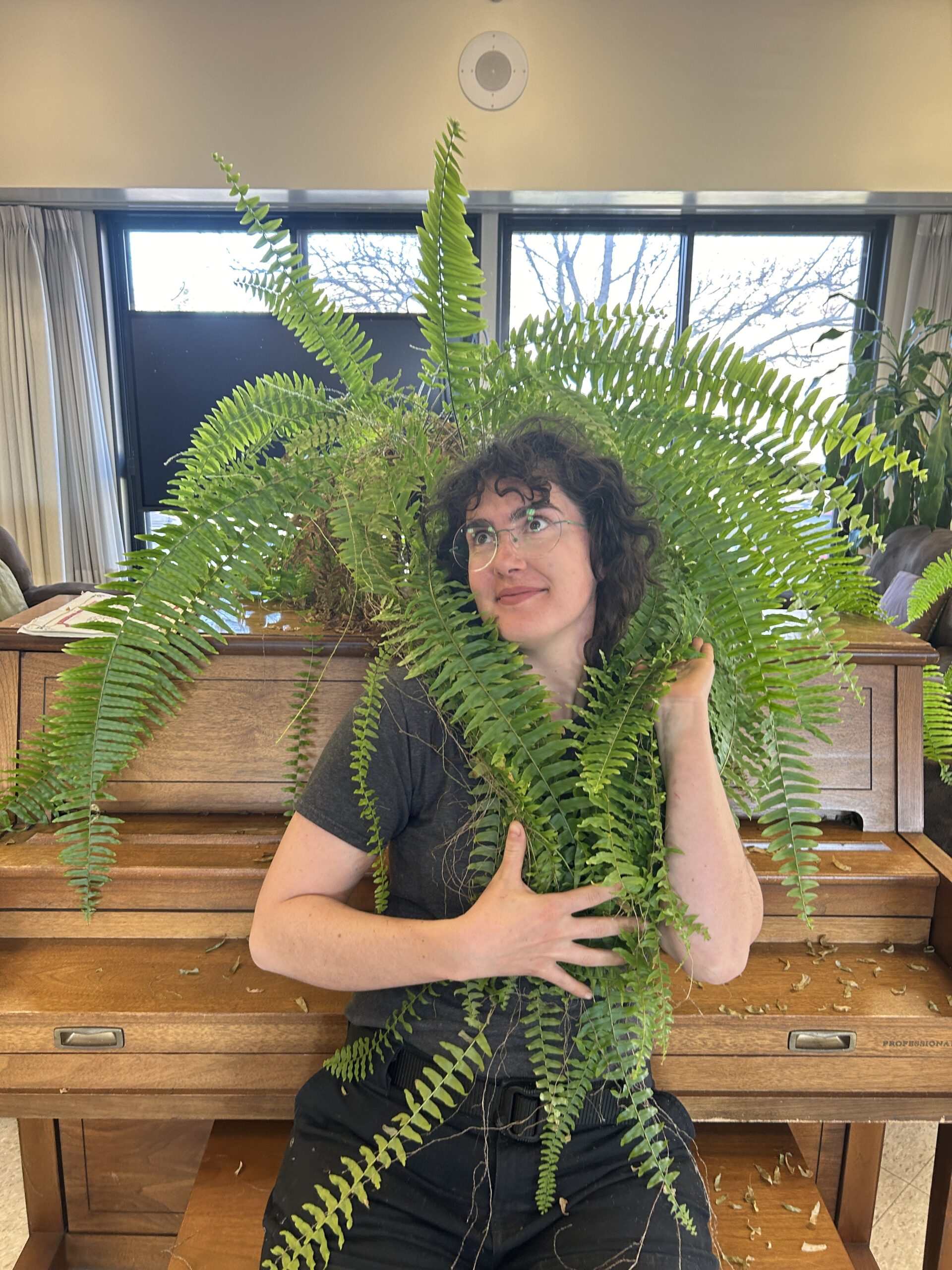 Starr is sitting on a bench in front of a piano. There is a large fern on top of the piano, and she is posing with the fronds around her shoulders like long hair.