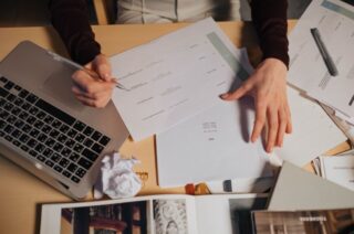 Stock image of a desk with a laptop, papers, and books. We see someone's hands as they read and write on a paper.