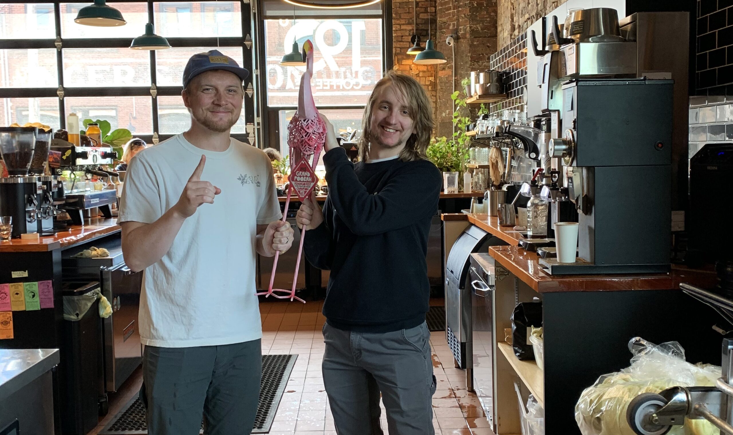 Two smiling people in a coffee shop hold up a large flamingo lawn ornament. One holds up a "number one" gesture.